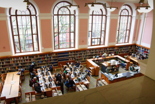 A spacious library with tall windows, bookshelves, and people using computers at study tables.