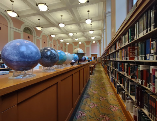 A library interior featuring a row of planetary models on a wooden table, with bookshelves lining the walls.