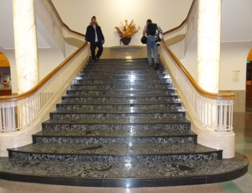 Two people walking down a grand staircase with decorative black marble steps and a floral arrangement at the top.