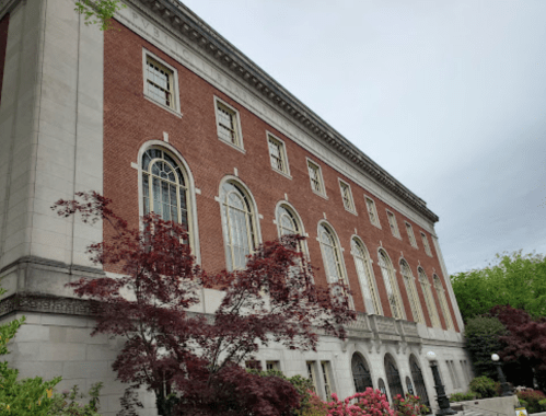 Historic brick building with large windows, surrounded by greenery and colorful shrubs under a cloudy sky.