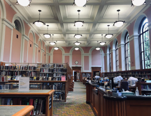 A spacious library interior with pink walls, tall windows, and bookshelves filled with books. Desks are arranged for study.