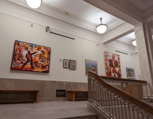 A gallery wall featuring colorful paintings, with a staircase and benches in a well-lit, elegant space.