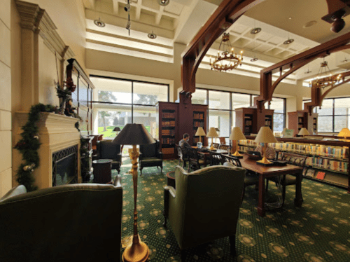 Cozy library interior with large windows, bookshelves, and comfortable seating, featuring a fireplace and lamps.