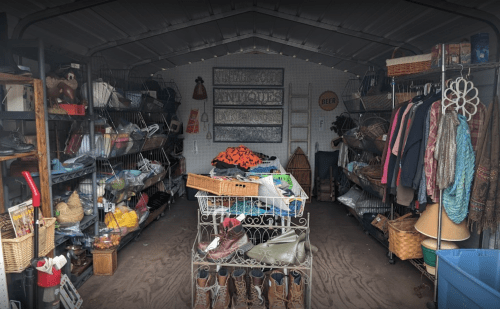 A cluttered thrift store interior with shelves of clothes, shoes, and various items in a shed-like space.