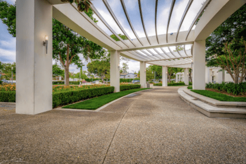 A curved pathway under a modern pergola, surrounded by greenery and trees in a landscaped area.