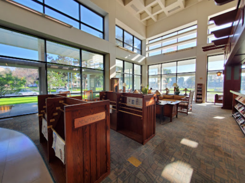 Bright library interior with large windows, wooden shelves, and reading areas, surrounded by greenery outside.