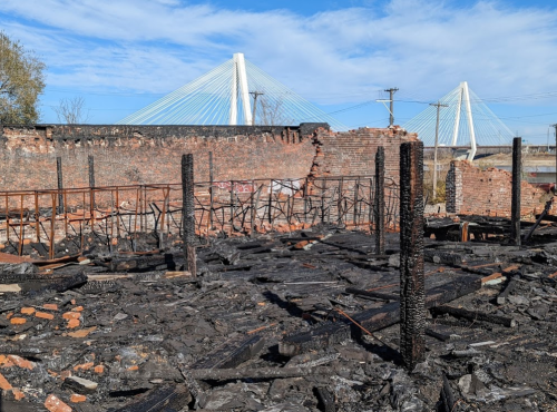 Burned building remnants with charred debris, against a backdrop of a bridge and blue sky.