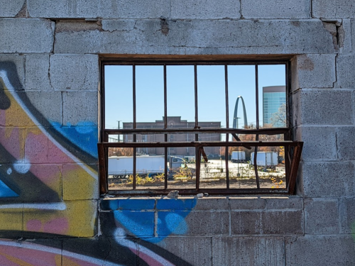 A broken window with a metal frame, revealing a view of a cityscape and colorful graffiti on the wall.