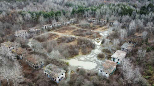 Aerial view of abandoned buildings surrounded by overgrown vegetation and a small pond in a desolate landscape.