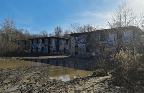Abandoned buildings covered in graffiti, surrounded by muddy ground and sparse trees under a clear blue sky.