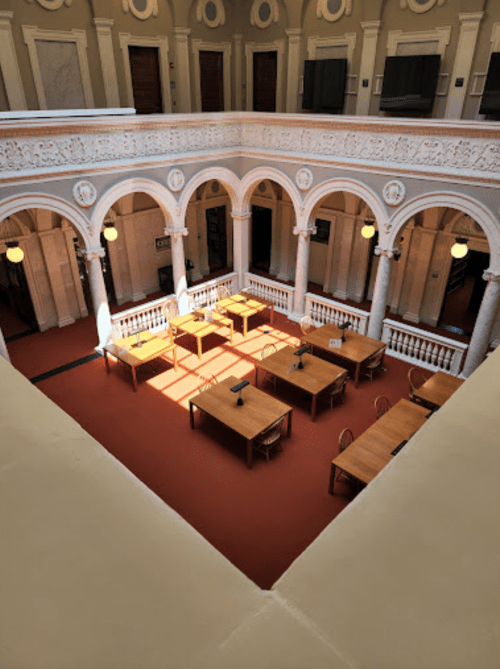 A spacious library interior with arched columns, wooden tables, and warm sunlight illuminating the red carpet.