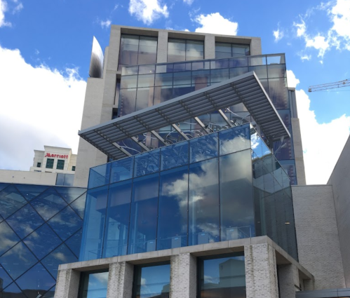 Modern building with glass facade and angular roof, reflecting clouds in the sky.