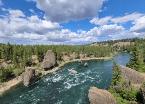 A scenic river flows through a forested landscape, with rocky formations and a blue sky dotted with clouds.