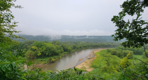 A misty river winding through lush green trees and hills under a cloudy sky.