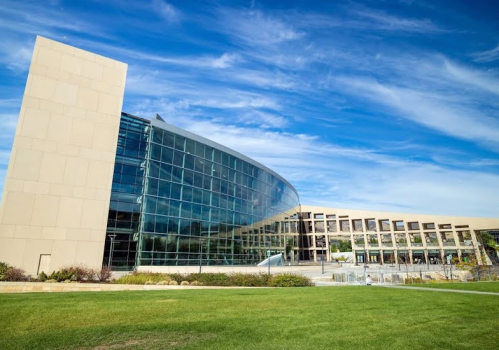 Modern building with large glass windows and a curved design, set against a blue sky and green lawn.