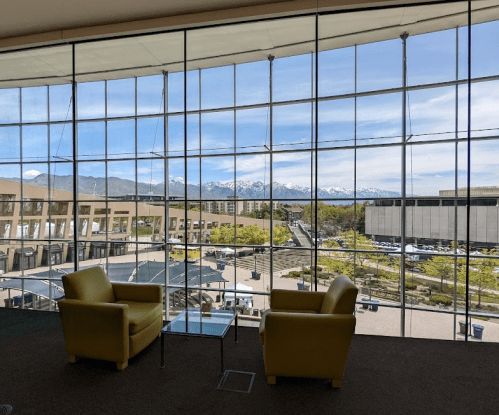 A spacious room with large windows showcasing a mountain view, featuring two yellow chairs and a glass table.