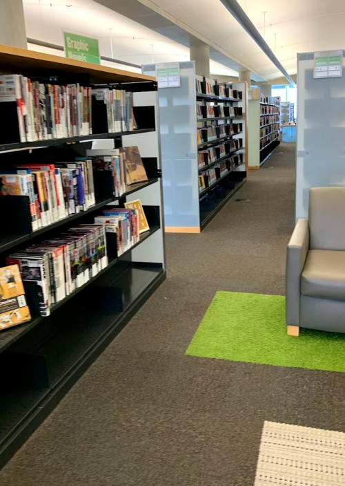 A library aisle with bookshelves on either side and a cozy seating area with a green rug.