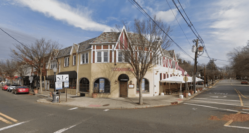 A charming corner building with a sloped roof, featuring outdoor seating and trees lining the street under a cloudy sky.