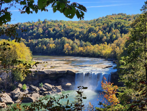 A scenic view of a waterfall surrounded by colorful autumn foliage and rocky terrain under a clear blue sky.