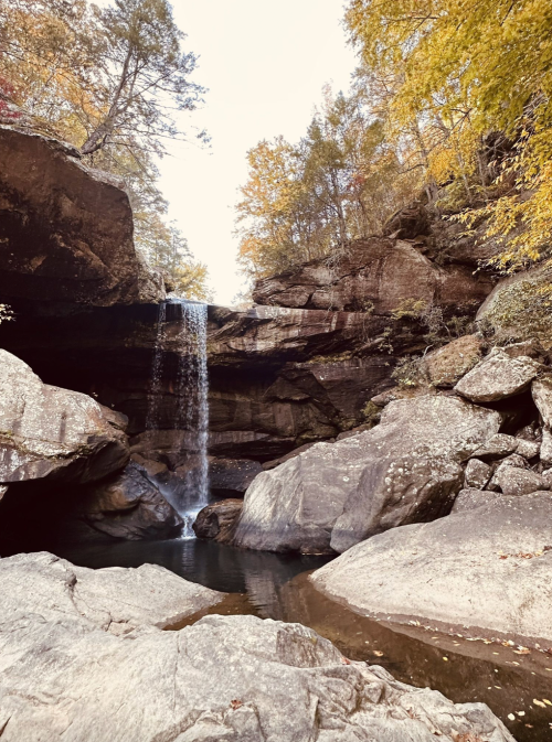 A serene waterfall cascades over rocks into a calm pool, surrounded by autumn foliage and rugged cliffs.
