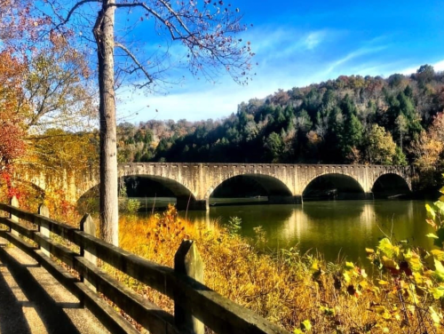 A scenic view of a stone bridge over a calm river, surrounded by autumn foliage and a clear blue sky.