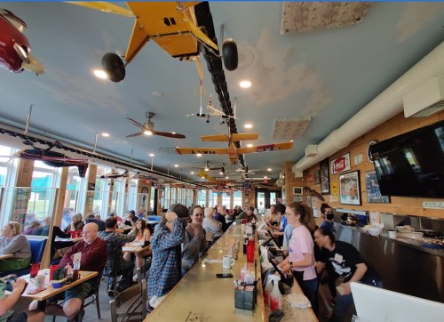 A busy restaurant interior with model airplanes hanging from the ceiling and patrons enjoying their meals at the counter.