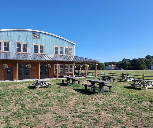 A rustic building with a curved roof, surrounded by picnic tables on a grassy field under a clear blue sky.