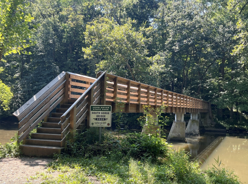 A wooden bridge spans a calm river, surrounded by lush green trees and a sign indicating the South Gorge Bridge.