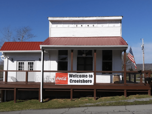 A white building with a red roof, featuring a "Welcome to Creeisboro" sign and an American flag.