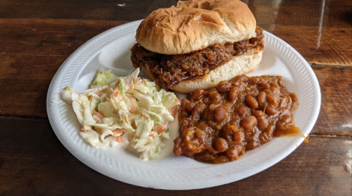 A plate with a pulled pork sandwich, baked beans, and coleslaw on a paper plate.
