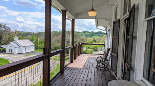 A porch with wooden flooring and rocking chairs, overlooking a scenic landscape with rolling hills and a small church.