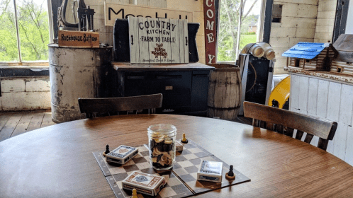 A rustic dining area with a wooden table, game pieces, and a country kitchen sign in a bright, airy space.