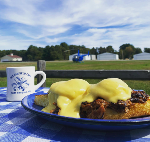 A plate of eggs Benedict with pulled pork, set on a checkered tablecloth, with a helicopter and hangars in the background.
