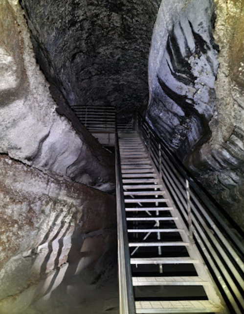 A dimly lit cave interior with a wooden staircase leading down, surrounded by rocky walls and shadows.