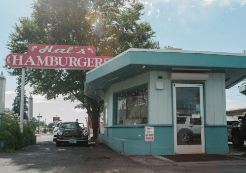 A retro hamburger stand with a large sign reading "Hal's Hamburgers" and a classic car parked outside.