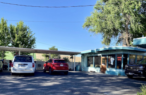 A small diner with a "DINE OPEN" sign, surrounded by parked cars and trees under a clear blue sky.