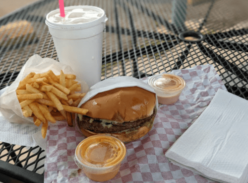 A burger with lettuce and pickles, fries, a drink, and two dipping sauces on a checkered paper on a table.