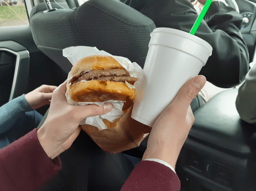 A hand holds a burger wrapped in paper, while another hand holds a white cup with a green straw inside a car.