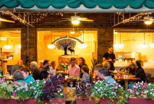 A bustling restaurant patio filled with diners enjoying meals, surrounded by flowers and warm lighting.