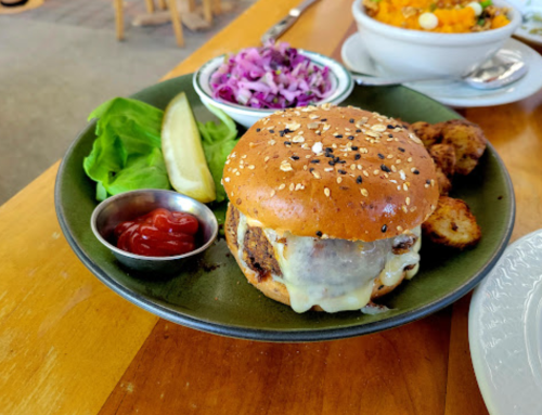 A sesame seed burger with cheese, lettuce, pickles, and ketchup, served with coleslaw and potato cakes on a green plate.