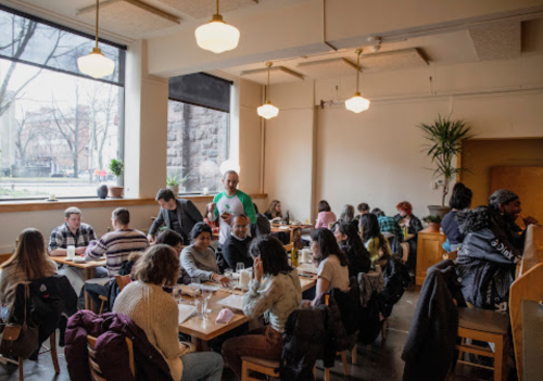 A busy café with people dining at wooden tables, large windows letting in natural light, and a relaxed atmosphere.