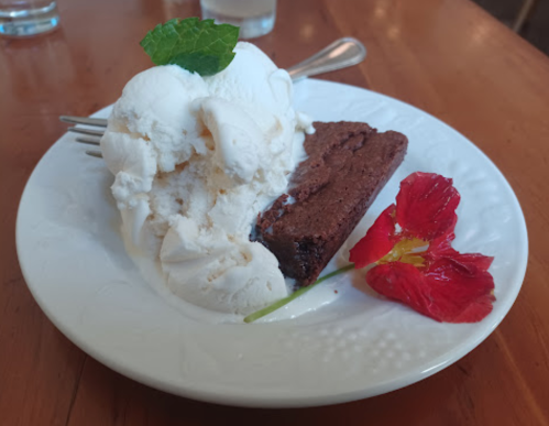 A plate with a slice of brownie, a scoop of vanilla ice cream, a mint leaf, and a flower garnish.