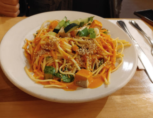 A plate of noodles topped with vegetables, tofu, sesame seeds, and a creamy sauce, served on a wooden table.