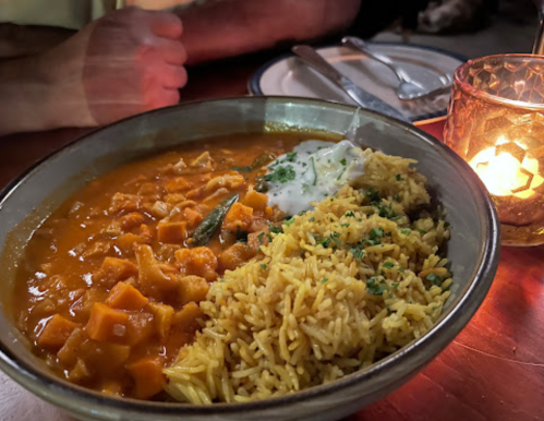 A bowl of curry with chunks of vegetables, served with fragrant rice and a side of yogurt, illuminated by candlelight.