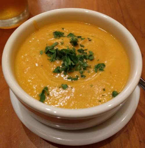 A bowl of creamy orange soup topped with fresh green herbs, served on a wooden table.