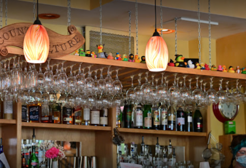 A bar with hanging glasses, colorful light fixtures, and rubber ducks lined along the top shelf.