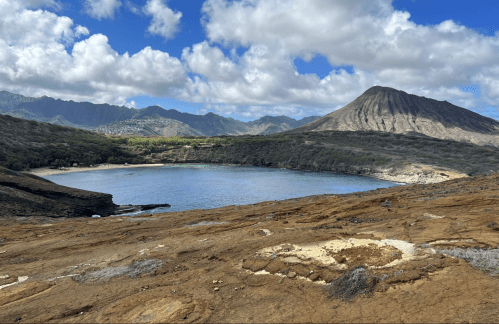A scenic view of a calm bay surrounded by rocky terrain and mountains under a partly cloudy sky.