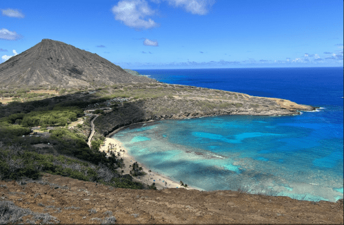 A scenic view of a coastline with turquoise waters, sandy beach, and a volcanic mountain in the background under a blue sky.