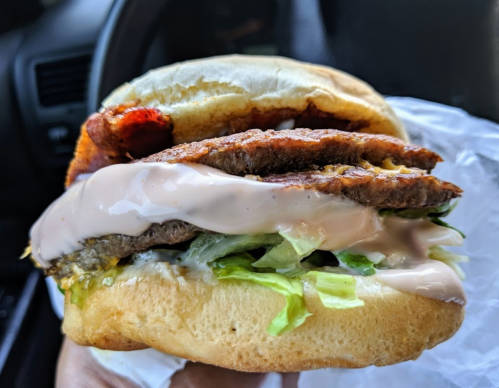 A close-up of a sandwich with layers of lettuce, sauce, and meat, held in a hand inside a car.