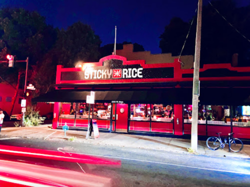 A brightly lit restaurant named "Sticky Rice" at night, with a red exterior and outdoor seating.